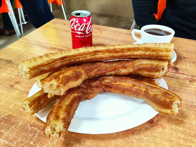 Plato blanco con varios churros largos y gruesos parcialmente mordidos, acompañado de una lata de Coca-Cola y una taza de chocolate caliente sobre una mesa de madera.